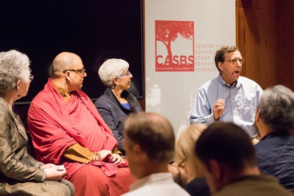 Three people wearing microphones, including a Buddhist monk, listen to a fourth person speak.