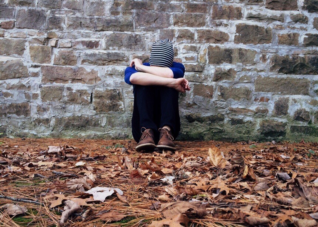 Woman sitting in leaves against a stone wall with her head down on her arms, folder over her knees